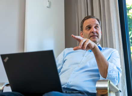 Geschäftsmann mit Laptop im Gespräch Ein Mann sitzt auf einem Stuhl und zeigt mit seiner Hand. Vor ihm steht ein Laptop. Er trägt ein hellblaues Hemd und hat einen nachdenklichen Gesichtsausdruck. Im Hintergrund sind Vorhänge und ein Fenster mit Blick ins Freie.
