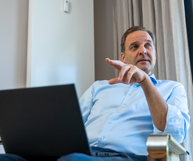 Geschäftsmann mit Laptop im Gespräch Ein Mann sitzt auf einem Stuhl und zeigt mit seiner Hand. Vor ihm steht ein Laptop. Er trägt ein hellblaues Hemd und hat einen nachdenklichen Gesichtsausdruck. Im Hintergrund sind Vorhänge und ein Fenster mit Blick ins Freie.