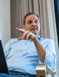 Geschäftsmann mit Laptop im Gespräch Ein Mann sitzt auf einem Stuhl und zeigt mit seiner Hand. Vor ihm steht ein Laptop. Er trägt ein hellblaues Hemd und hat einen nachdenklichen Gesichtsausdruck. Im Hintergrund sind Vorhänge und ein Fenster mit Blick ins Freie.