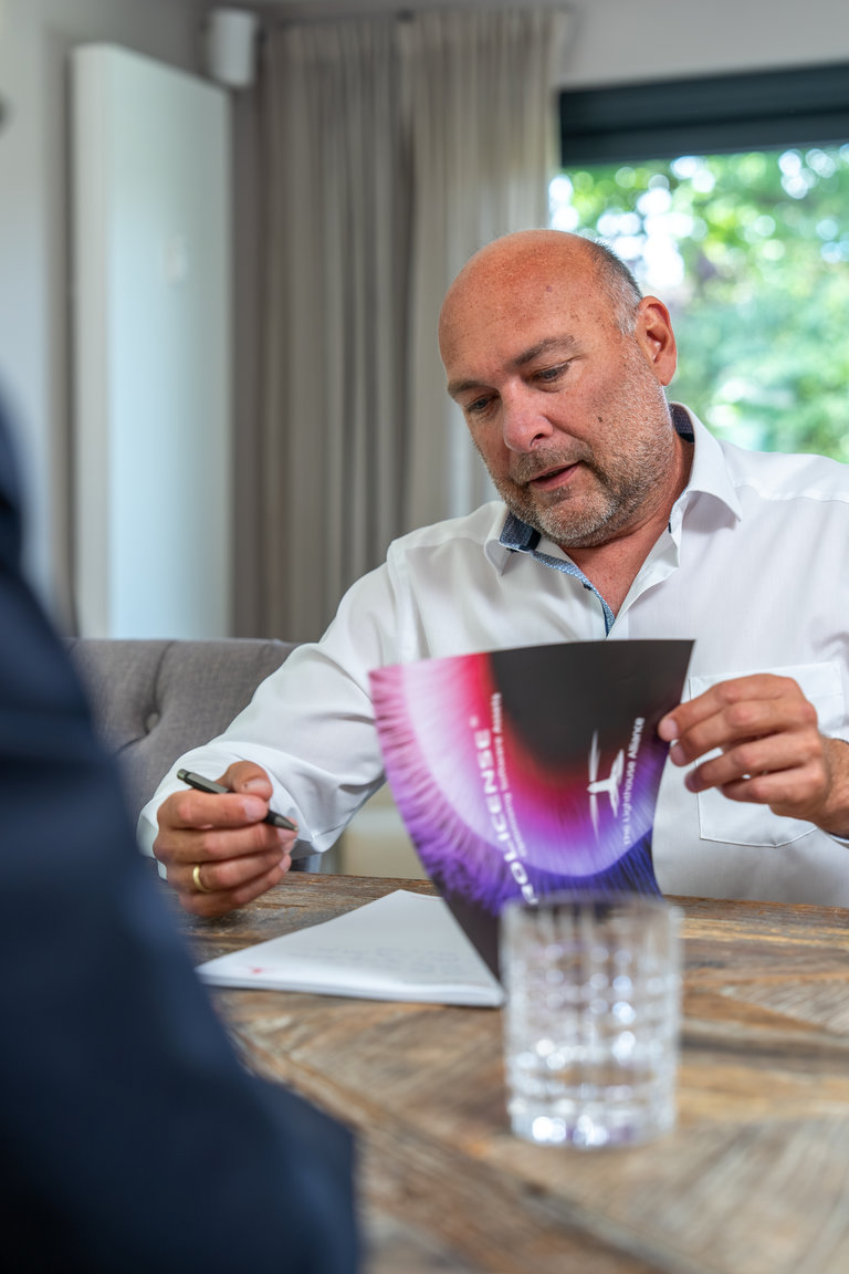 Geschäftliche Besprechung am Tisch Ein Mann mit Glatze und Bart sitzt an einem Tisch und hält ein farbiges Dokument in der Hand. Er trägt ein weißes Hemd und schreibt mit einem Stift auf einem Blatt Papier. Im Hintergrund sind ein Fenster und Vorhänge sichtbar.