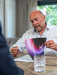 Geschäftliche Besprechung am Tisch Ein Mann mit Glatze und Bart sitzt an einem Tisch und hält ein farbiges Dokument in der Hand. Er trägt ein weißes Hemd und schreibt mit einem Stift auf einem Blatt Papier. Im Hintergrund sind ein Fenster und Vorhänge sichtbar.