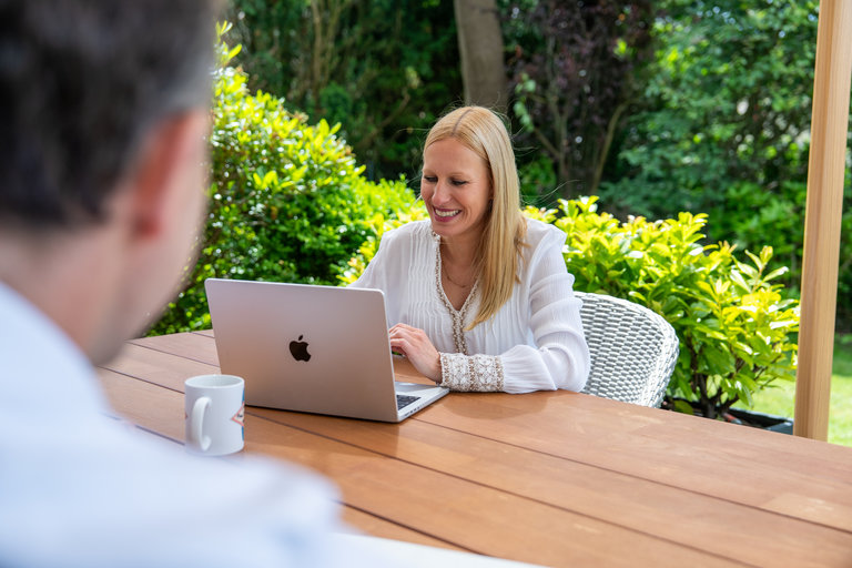 Frau arbeitet am Laptop im Freien Eine Frau sitzt an einem Tisch im Freien und lächelt, während sie auf ihrem Laptop arbeitet. Im Vordergrund ist ein unscharfes Bild eines Mannes zu sehen, der einen Kaffeebecher hält. Um sie herum sind grüne Pflanzen und ein sonniger Garten.