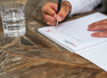 Eine Hand hält einen Stift und schreibt auf ein Blatt Papier, das auf einem Tisch liegt. Neben dem Blatt steht ein Glas Wasser. Der Tisch hat eine holzähnliche Struktur.