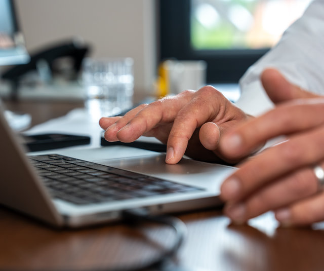 Arbeiten am Laptop im Büro Hände eines Mannes, die auf einer Laptop-Tastatur tippen. Im Hintergrund sind ein Glas Wasser und ein Smartphone sichtbar. Der Arbeitsplatz ist hell und modern eingerichtet.