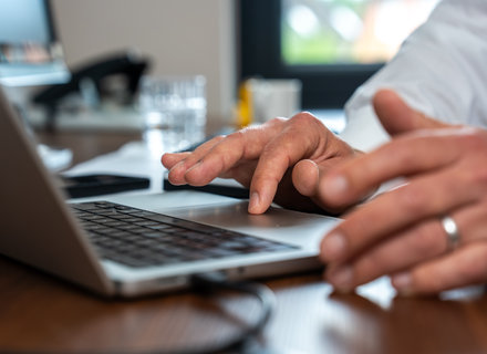Hände eines Mannes, die auf einer Laptop-Tastatur tippen. Im Hintergrund sind ein Glas Wasser und ein Smartphone sichtbar. Der Arbeitsplatz ist hell und modern eingerichtet.