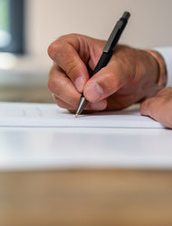 Hand schreibt in Notizbuch Eine Hand hält einen Stift und schreibt in ein Notizbuch, das auf einem Holztisch liegt. Im Hintergrund ist ein Wasserglas und ein Fenster zu sehen.