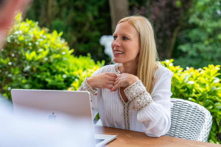 Geschäftliches Treffen im Freien Eine lächelnde Frau sitzt an einem Tisch im Freien, wo sie mit einem Laptop arbeitet. Im Hintergrund sind grüne Pflanzen zu sehen. Die Frau trägt ein weißes Oberteil mit dekorativen Elementen und sieht interessiert aus, während sie gestikuliert.
