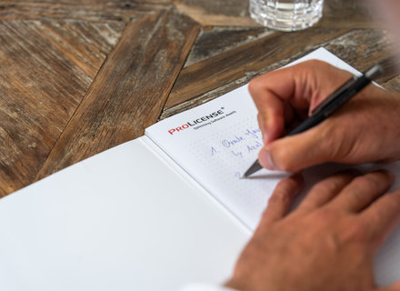 Notizen auf einem Notizblock Eine Hand schreibt mit einem Stift auf ein Blatt Papier in einem Notizbuch. Das Papier zeigt Notizen und ist auf einem Tisch mit einer holzartigen Oberfläche platziert. Im Hintergrund ist ein Glas sichtbar.