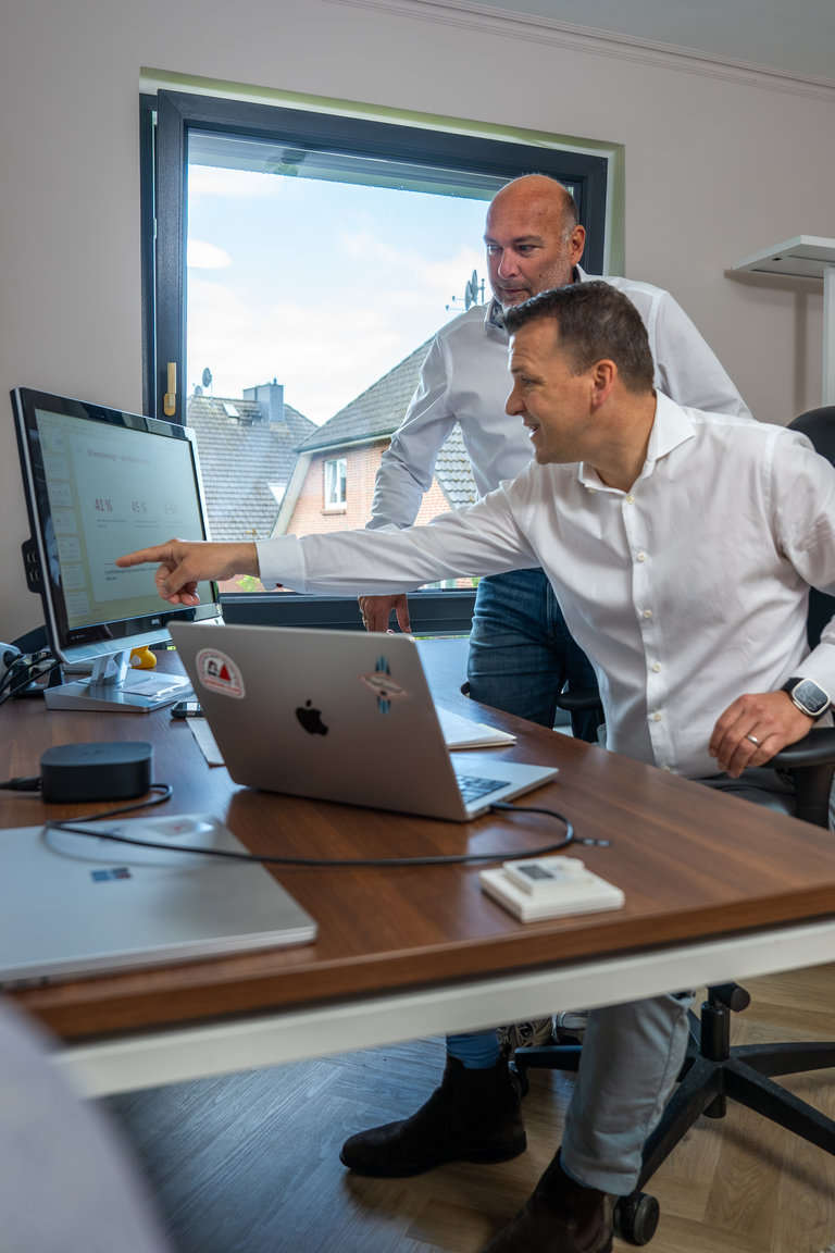 Teamarbeit am Computer Arbeitsplatz Zwei Männer stehen in einem modernen Büro vor einem Computer. Der Mann im Vordergrund zeigt auf den Bildschirm, während der andere hinten interessiert zuschaut. Auf dem Tisch sind Laptops und ein kleiner Monitor zu sehen, und ein Fenster lässt Tageslicht herein.