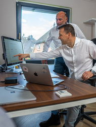 Teamarbeit am Computer Arbeitsplatz Zwei Männer stehen in einem modernen Büro vor einem Computer. Der Mann im Vordergrund zeigt auf den Bildschirm, während der andere hinten interessiert zuschaut. Auf dem Tisch sind Laptops und ein kleiner Monitor zu sehen, und ein Fenster lässt Tageslicht herein.