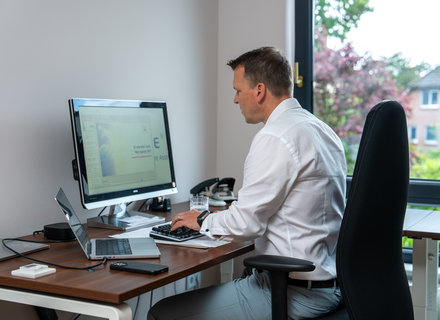 Heimarbeitsplatz im modernen Büro Ein Mann in einem weißen Hemd sitzt an einem Schreibtisch und arbeitet an einem Computer. Der Bildschirm zeigt eine Präsentation. Auf dem Tisch befinden sich ein Laptop, Schreibmaterial und ein Glas Wasser. Im Hintergrund sind Fenster und eine grüne Aussicht zu sehen.