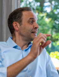 Gespräch in hellem Raum Ein Mann sitzt auf einem Sofa und gestikuliert mit einer Hand. Er trägt ein hellblaues Hemd und schaut zur Seite. Im Hintergrund sind verschwommen Bäume und Sitzgelegenheiten im Freien zu sehen.