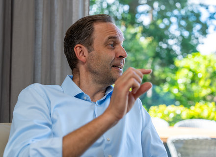 Ein Mann sitzt auf einem Sofa und gestikuliert mit einer Hand. Er trägt ein hellblaues Hemd und schaut zur Seite. Im Hintergrund sind verschwommen Bäume und Sitzgelegenheiten im Freien zu sehen.