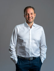 Lächelnder Mann im Hemd Ein lächelnder Mann steht mit den Händen in den Taschen da. Er trägt ein weißes Hemd und eine dunkle Hose. Der Hintergrund ist grau und neutral.