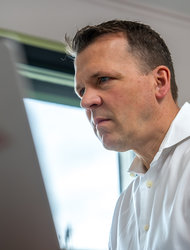 Fokusierter Mann am Laptop Ein Mann in einem weißen Hemd arbeitet konzentriert an einem Laptop. Er sitzt an einem Tisch, wobei im Hintergrund Fenster mit Tageslicht zu sehen sind.