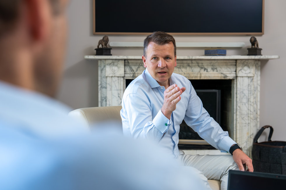 Gespräch in modernem Ambiente Ein Mann in einem hellblauen Hemd sitzt auf einem Sofa und spricht gestikulierend. Im Hintergrund ist ein Kamin und ein Fernseher. Die Perspektive zeigt teilweise eine andere Person, die ihm gegenüber sitzt.