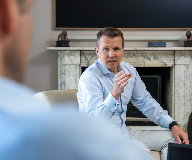 Gespräch in modernem Ambiente Ein Mann in einem hellblauen Hemd sitzt auf einem Sofa und spricht gestikulierend. Im Hintergrund ist ein Kamin und ein Fernseher. Die Perspektive zeigt teilweise eine andere Person, die ihm gegenüber sitzt.