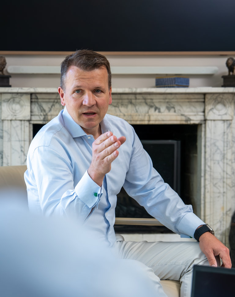 Gespräch in modernem Ambiente Ein Mann in einem hellblauen Hemd sitzt auf einem Sofa und spricht gestikulierend. Im Hintergrund ist ein Kamin und ein Fernseher. Die Perspektive zeigt teilweise eine andere Person, die ihm gegenüber sitzt.