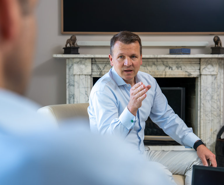 Gespräch in modernem Ambiente Ein Mann in einem hellblauen Hemd sitzt auf einem Sofa und spricht gestikulierend. Im Hintergrund ist ein Kamin und ein Fernseher. Die Perspektive zeigt teilweise eine andere Person, die ihm gegenüber sitzt.