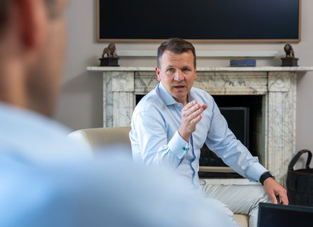 Gespräch in modernem Ambiente Ein Mann in einem hellblauen Hemd sitzt auf einem Sofa und spricht gestikulierend. Im Hintergrund ist ein Kamin und ein Fernseher. Die Perspektive zeigt teilweise eine andere Person, die ihm gegenüber sitzt.