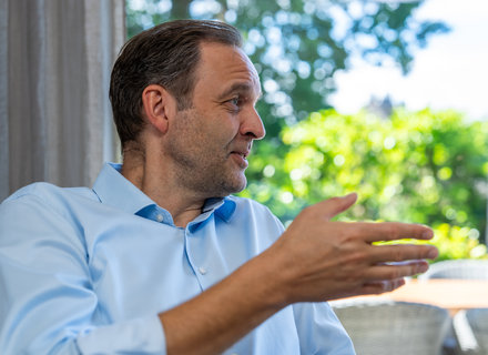 Gespräch im Freien Ein Mann mit kurzen Haaren und in einem hellblauen Hemd sitzt im Gespräch. Er gestikuliert mit der rechten Hand und hat einen nachdenklichen Gesichtsausdruck. Der Hintergrund zeigt grüne Pflanzen und sonniges Wetter.