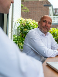 Gespräch im Freien bei Tageslicht Zwei Männer sitzen an einem Tisch im Freien. Einer der Männer, im weißen Hemd, blickt lächelnd in die Kamera. Auf dem Tisch steht eine Tasse und ein Laptop. Im Hintergrund sind grüne Pflanzen zu sehen und ein Gebäude.