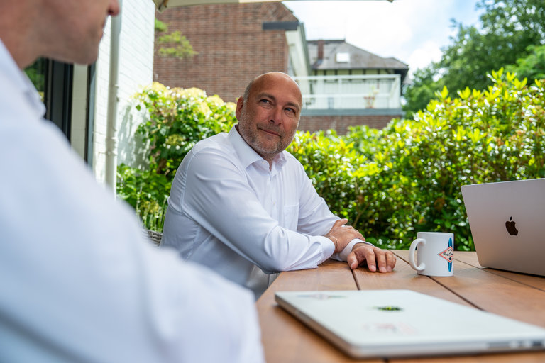 Gespräch im Freien bei Tageslicht Zwei Männer sitzen an einem Tisch im Freien. Einer der Männer, im weißen Hemd, blickt lächelnd in die Kamera. Auf dem Tisch steht eine Tasse und ein Laptop. Im Hintergrund sind grüne Pflanzen zu sehen und ein Gebäude.