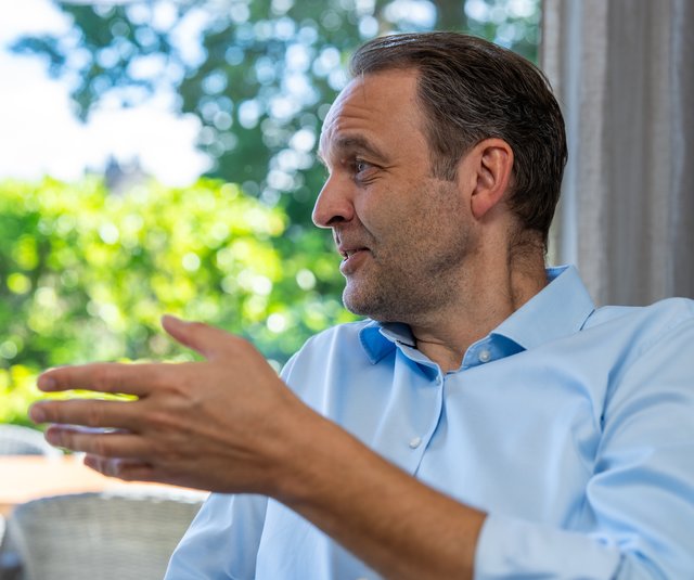 Ein Mann in einem hellblauen Hemd spricht nachdenklich. Er ist seitlich zu sehen, während er mit einer Hand gestikuliert. Im Hintergrund sind grüne Bäume und ein heller Raum erkennbar.