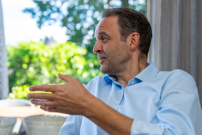 Ein Mann in einem hellblauen Hemd spricht nachdenklich. Er ist seitlich zu sehen, während er mit einer Hand gestikuliert. Im Hintergrund sind grüne Bäume und ein heller Raum erkennbar.