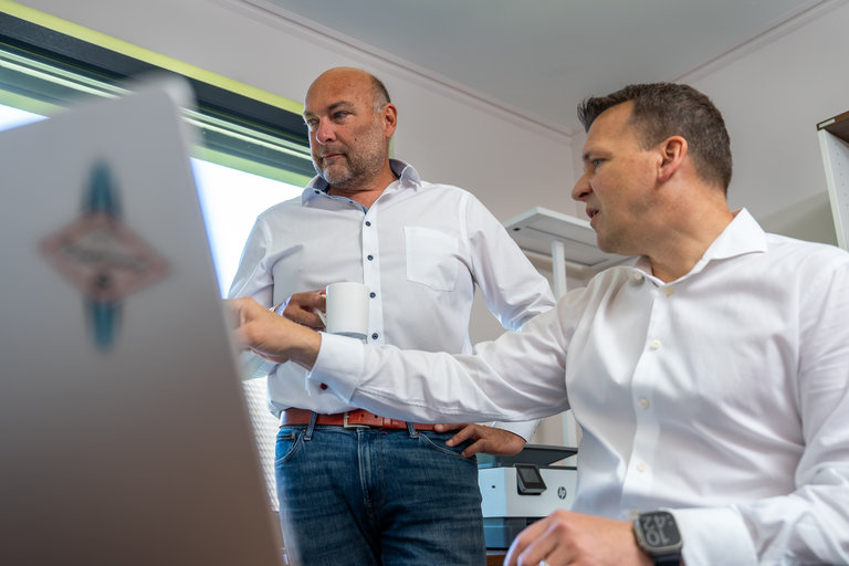 Geschäftliche Besprechung am Schreibtisch Zwei Männer in einem Büro stehen und diskutieren. Der Mann links hält eine Tasse, während der Mann rechts auf einen Laptopbildschirm zeigt. Beide tragen weiße Hemden und wirken engagiert in ihrem Gespräch.