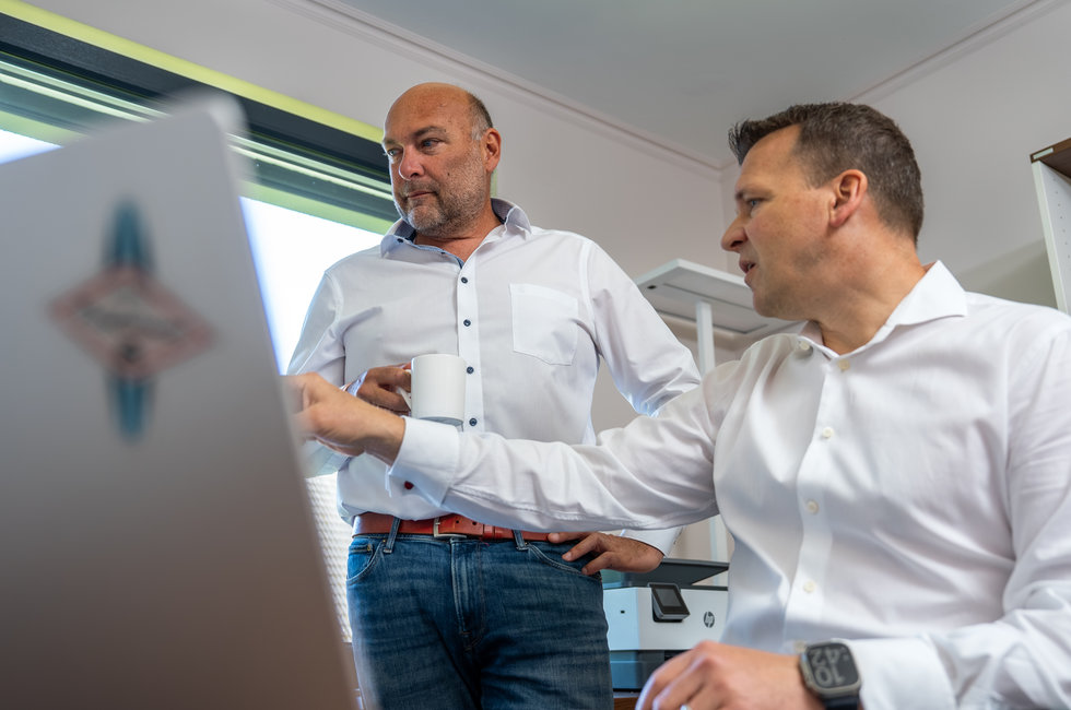 Geschäftliche Besprechung am Schreibtisch Zwei Männer in einem Büro stehen und diskutieren. Der Mann links hält eine Tasse, während der Mann rechts auf einen Laptopbildschirm zeigt. Beide tragen weiße Hemden und wirken engagiert in ihrem Gespräch.