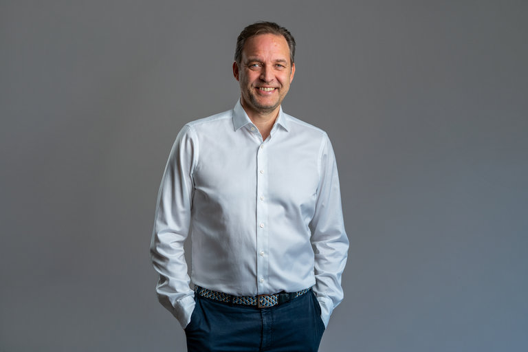 Professionelles Porträt eines Mannes Ein Mann steht mit einem Lächeln und lässig mit den Händen in den Taschen vor einem grauen Hintergrund. Er trägt ein weißes Hemd und dunkle Hose.