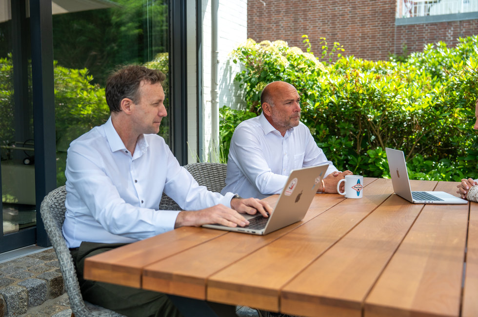 Geschäftliches Meeting im Freien Zwei Männer sitzen an einem Tisch im Freien. Der Mann links arbeitet an einem Laptop, während der Mann rechts auf einen Laptop und eine Tasse schaut. Im Hintergrund sind grüne Pflanzen zu sehen.