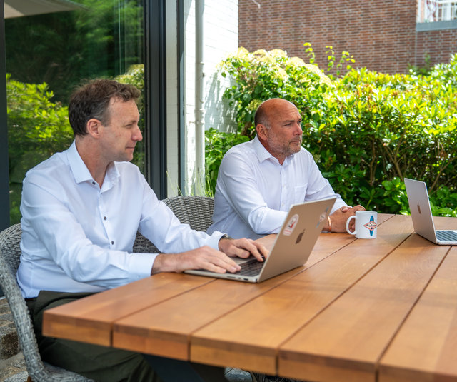 Geschäftliches Meeting im Freien Zwei Männer sitzen an einem Tisch im Freien. Der Mann links arbeitet an einem Laptop, während der Mann rechts auf einen Laptop und eine Tasse schaut. Im Hintergrund sind grüne Pflanzen zu sehen.