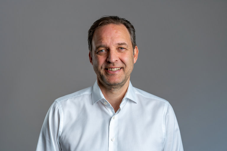 Porträt eines lächelnden Mannes Ein Mann mittleren Alters mit kurzen, braunen Haaren und einem freundlichen Lächeln steht vor einem grauen Hintergrund. Er trägt ein weißes Hemd mit offenen Kragen. Die Haltung ist selbstbewusst und einladend.