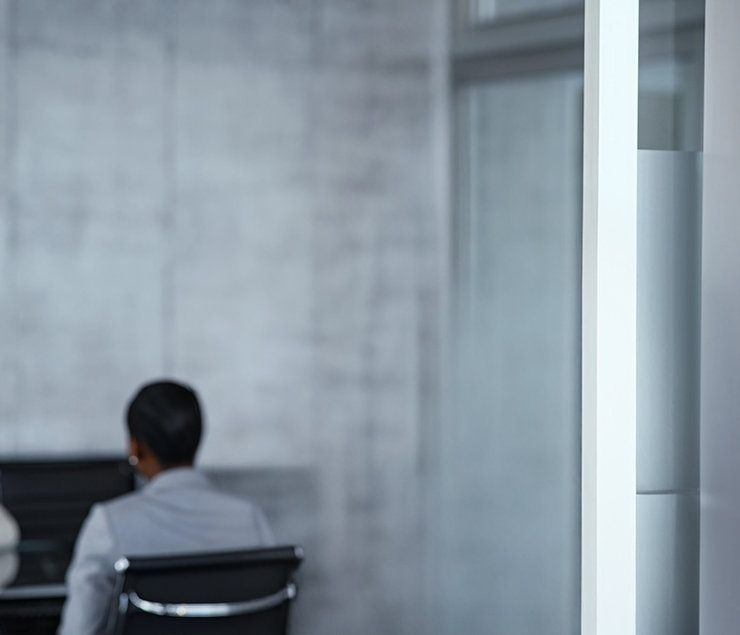 Besprechung im modernen Büro Zwei Personen sitzen in einem modernen Besprechungsraum. Eine Person, die im Anzug gekleidet ist, sitzt mit dem Rücken zur Kamera. Im Hintergrund ist eine weitere Person zu sehen, ebenfalls in formeller Kleidung. Der Raum hat eine minimalistische Gestaltung mit einem hellen, offenen Ambiente.