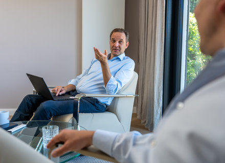 Gespräch in modernem Büro Zwei Männer führen ein Gespräch in einem hellen Raum. Einer sitzt mit einem Laptop auf einem Sessel, gestikuliert mit der Hand und schaut den anderen an, der leicht abgewandt ist. Ein Tisch mit einem Glas Wasser steht zwischen ihnen.
