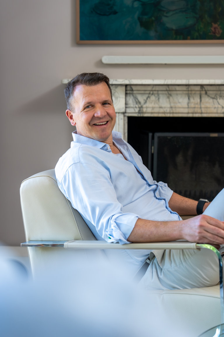 Lächelnder Mann im Wohnzimmer Ein lächelnder Mann sitzt entspannt in einem modernen Sessel. Er trägt ein hellblaues Hemd und hat kurze, dunkle Haare. Im Hintergrund ist ein Kamin und ein Gemälde sichtbar. Der Mann strahlt Freundlichkeit und Gelassenheit aus.