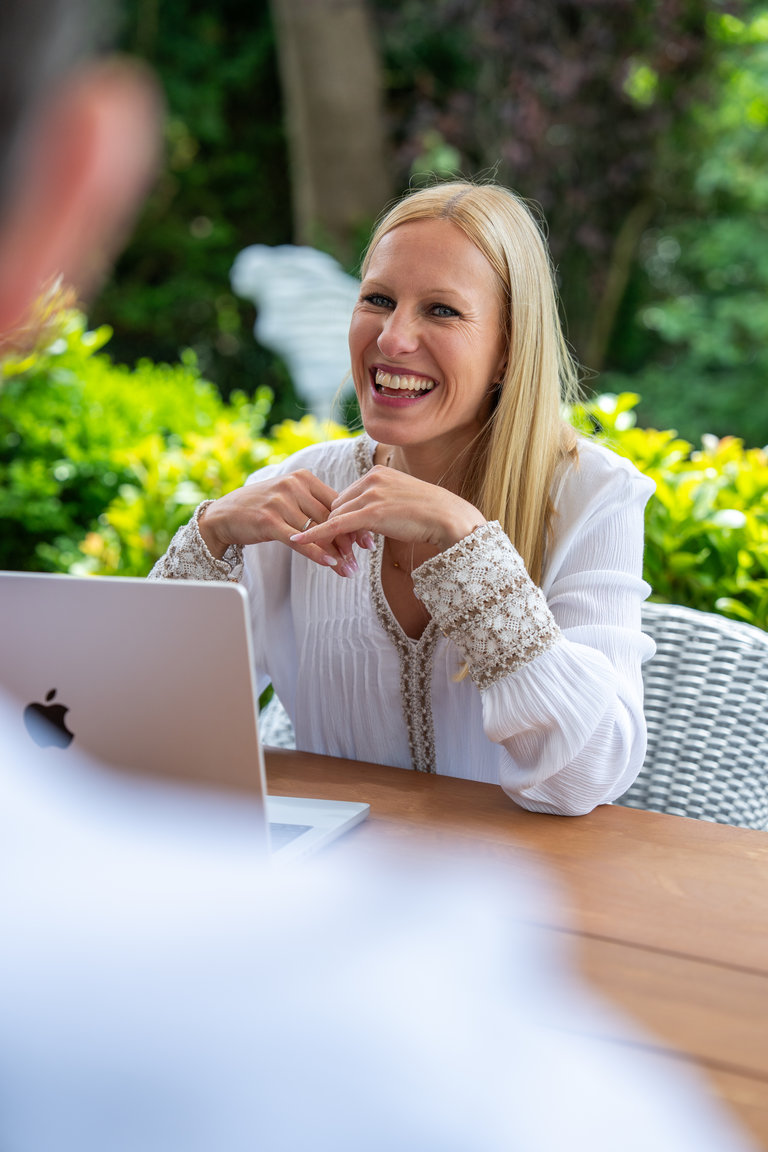 Lächelnde Frau am Tisch Eine lächelnde Frau mit langen blonden Haaren sitzt an einem Tisch im Freien, während sie mit einer Person spricht. Vor ihr steht ein Laptop, und im Hintergrund sind grüne Bäume und Pflanzen zu sehen.