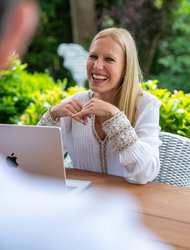 Lächelnde Frau am Tisch Eine lächelnde Frau mit langen blonden Haaren sitzt an einem Tisch im Freien, während sie mit einer Person spricht. Vor ihr steht ein Laptop, und im Hintergrund sind grüne Bäume und Pflanzen zu sehen.
