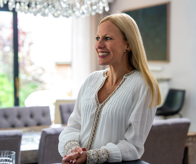 Lächelnde Frau im modernen Raum Eine Frau mit langen, blonden Haaren trägt ein weißes Oberteil und lächelt, während sie in einem modernen, hellen Raum steht. Im Hintergrund sind Möbel und Fenster sichtbar, durch die Tageslicht strömt.