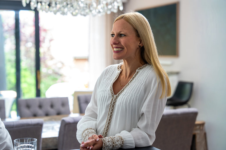Lächelnde Frau im modernen Raum Eine Frau mit langen, blonden Haaren trägt ein weißes Oberteil und lächelt, während sie in einem modernen, hellen Raum steht. Im Hintergrund sind Möbel und Fenster sichtbar, durch die Tageslicht strömt.