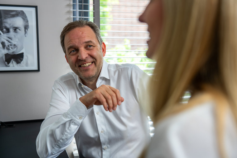 Gespräch in einem Büro Ein Mann in einem weißen Hemd sitzt an einem Tisch und lächelt, während er mit einer Frau spricht, die nicht vollständig sichtbar ist. Im Hintergrund ist ein Schwarz-Weiß-Foto eines Mannes zu sehen, und es gibt Fenster mit Jalousien, die Licht hereinlassen.