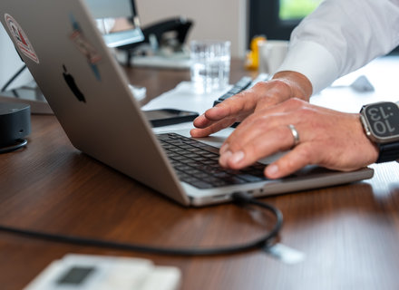Arbeiten am Laptop im Büro Eine Person tippt auf der Tastatur eines MacBook-Laptops. An einem Holztisch sind weitere Bürogegenstände und ein Wasserglas zu sehen. Ein Smartwatch ist am Handgelenk der Person zu erkennen.