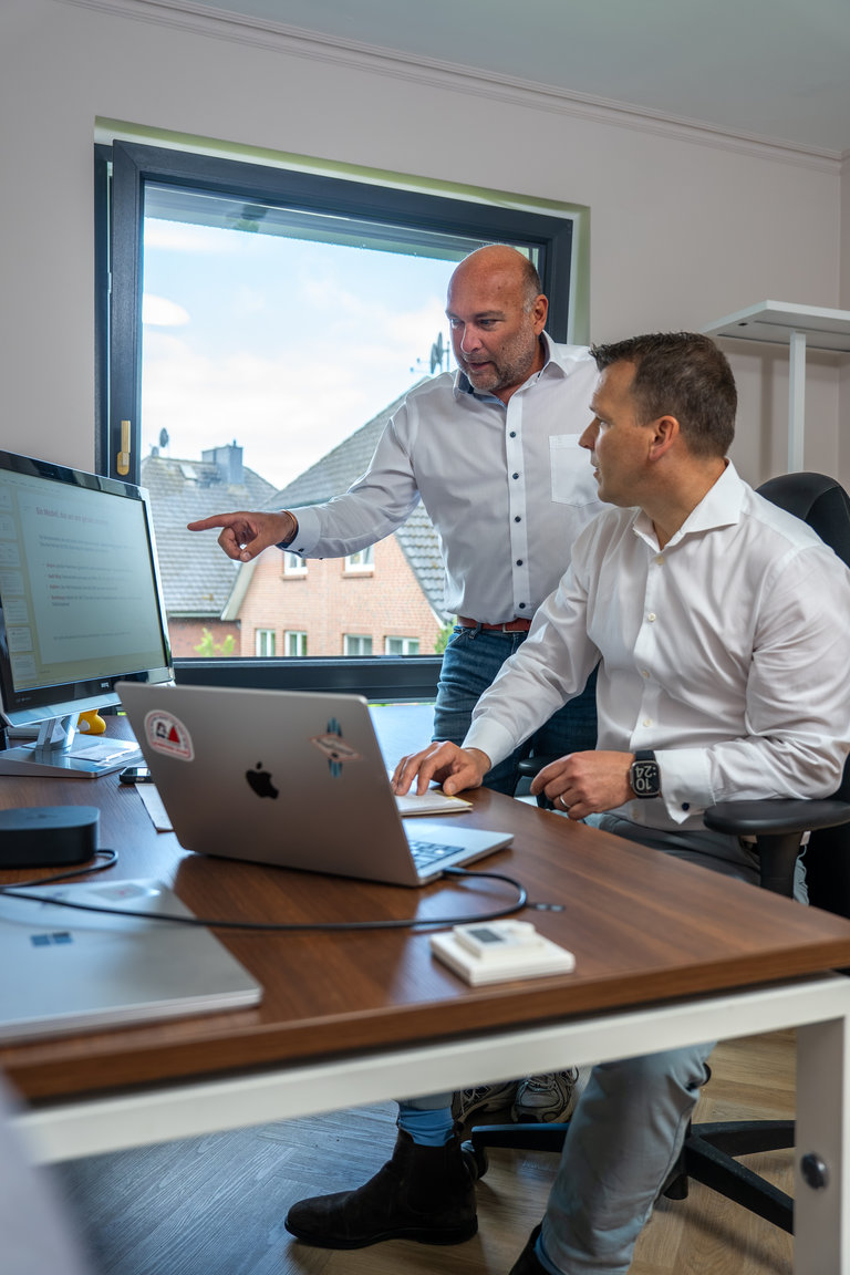 Teamarbeit im modernen Büro Zwei Männer stehen an einem Schreibtisch mit zwei Computern. Der Mann links weist auf den Bildschirm, während der andere sitzt und zuhört. Der Raum hat ein Fenster mit Blick auf ein Gebäude im Freien.